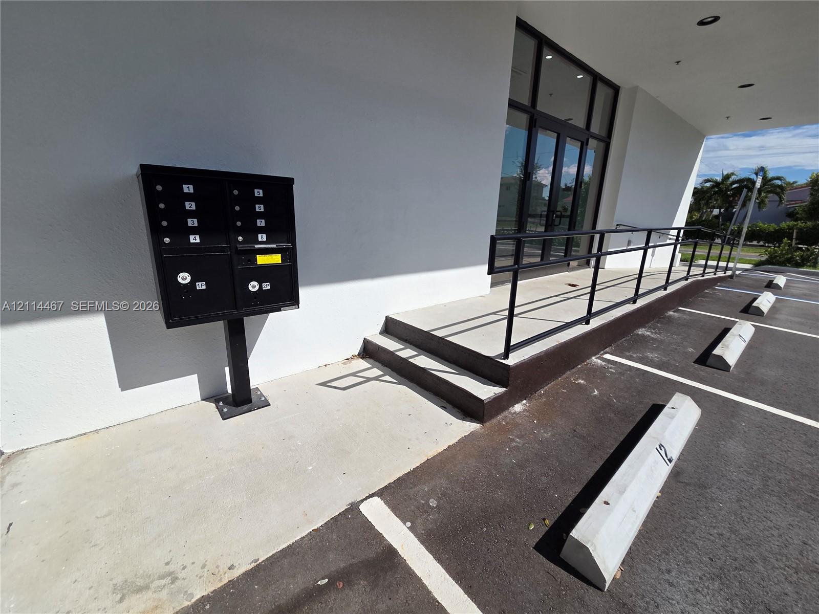 Exterior entrance featuring a concrete ramp with black metal handrails, a multi-unit wall-mounted mailbox system, and marked parking spaces with concrete wheel stops