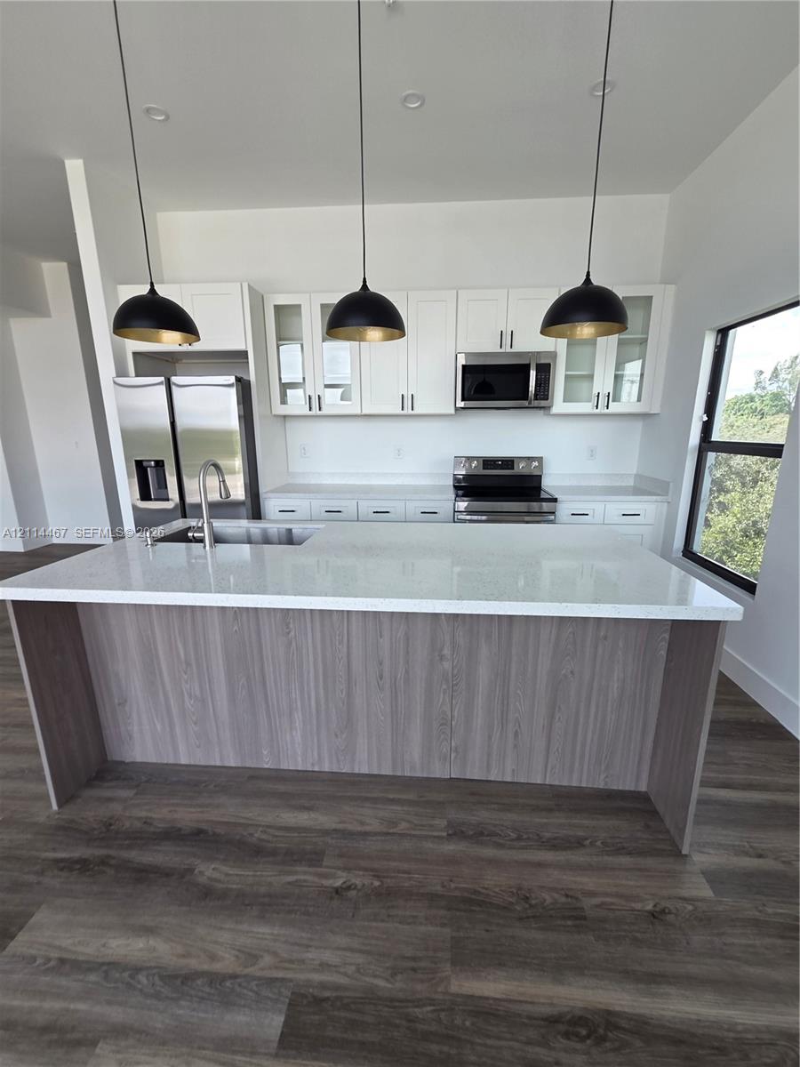 Kitchen featuring a center island with a stone-surface countertop, white shaker-style cabinetry, stainless steel appliances, wood-finish flooring, and three oversized black pendant lights