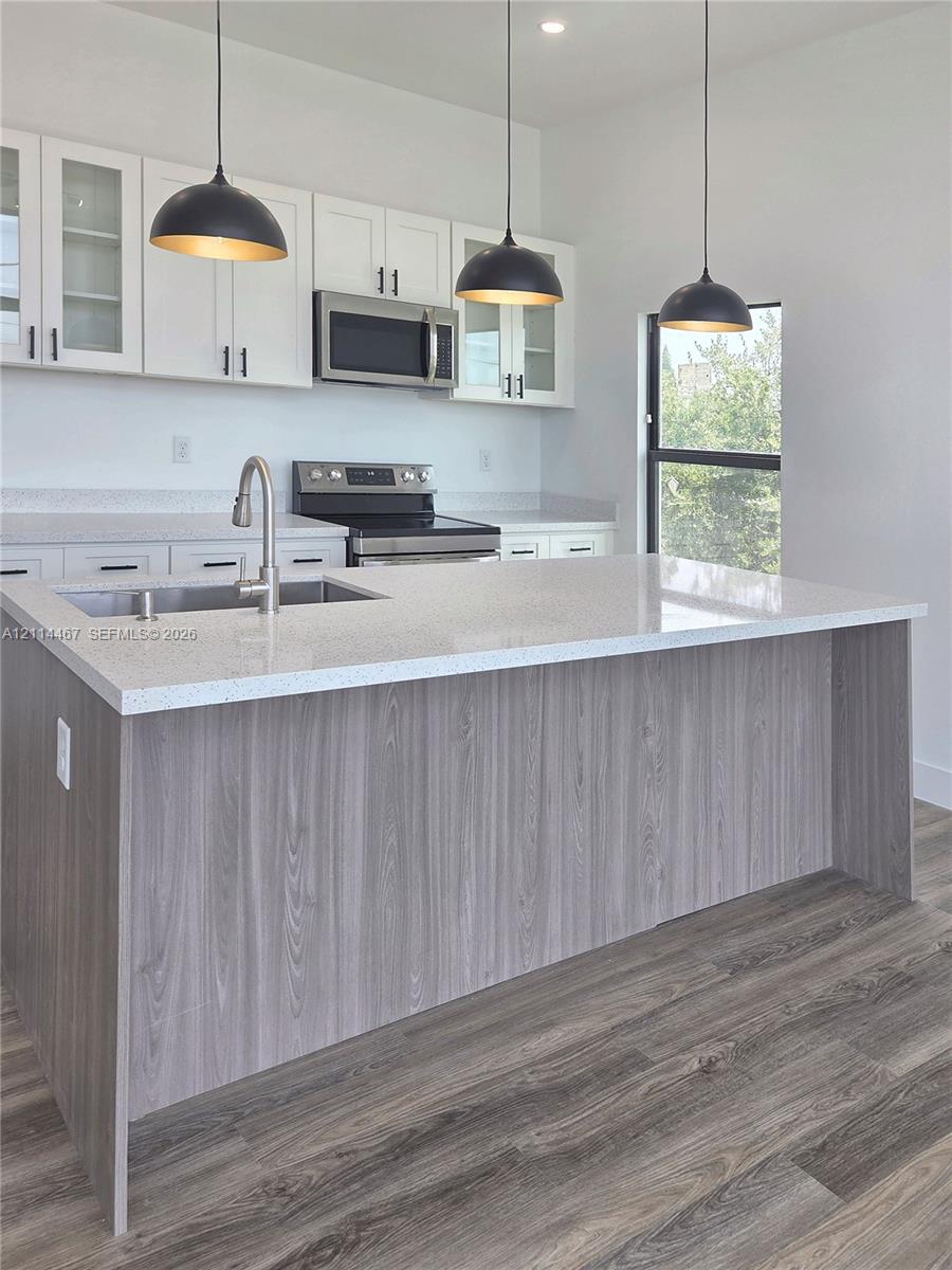 Kitchen featuring an island with a light-toned solid surface countertop and wood-finish cabinetry, white shaker-style upper cabinets, wood-finish flooring, and three matte black pendant lights