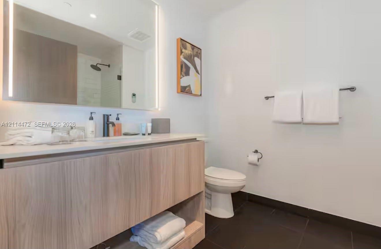 Floating vanity with wood-finish cabinetry and light-toned countertop, large rectangular mirror with integrated LED lighting, matte black fixtures, and dark-toned floor tile