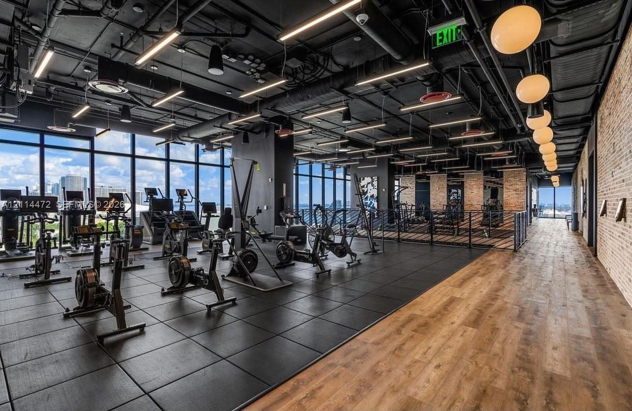 Fitness center featuring floor-to-ceiling windows, exposed industrial ceilings with recessed lighting, wood-finish flooring, and a textured brick accent wall