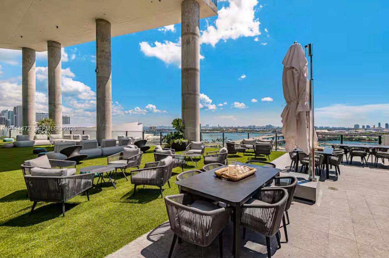 Expansive rooftop terrace featuring synthetic turf flooring, stone tile decking, structural concrete columns, and a glass panel perimeter railing