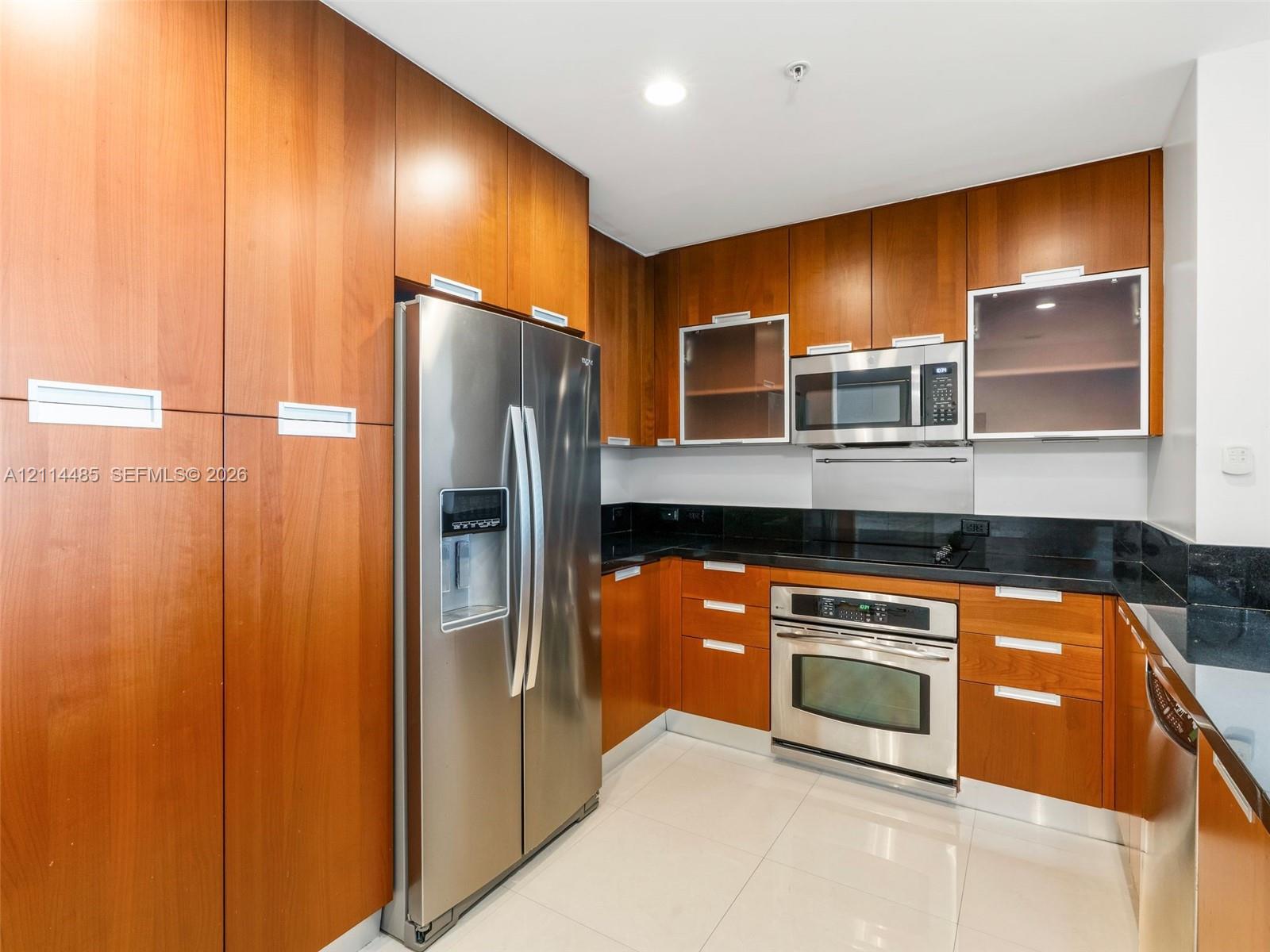 Kitchen featuring full-height cherry-finish cabinetry, stainless steel appliances, dark stone countertops, upper cabinets with glass-panel inserts, and light-tone tile flooring