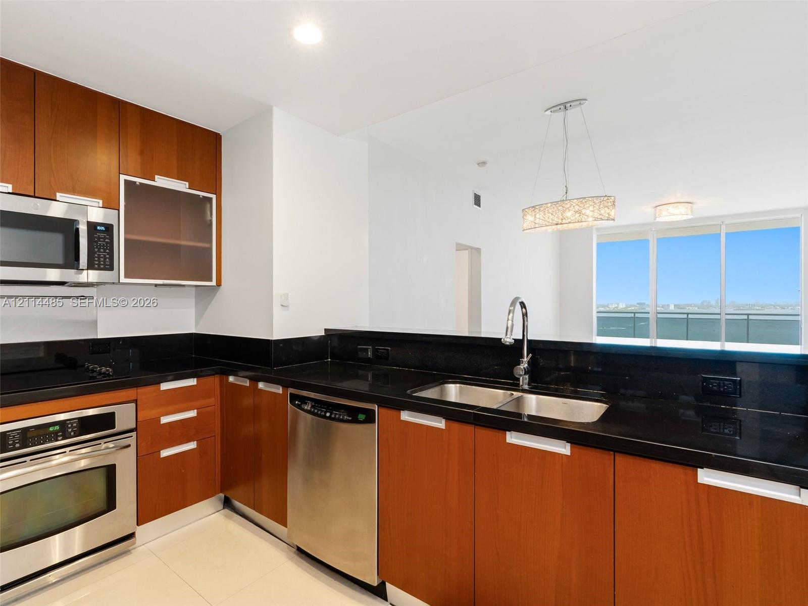 Kitchen featuring wood-finish cabinetry, dark stone countertops, stainless steel appliances, dual-basin sink with gooseneck faucet, and integrated pendant lighting