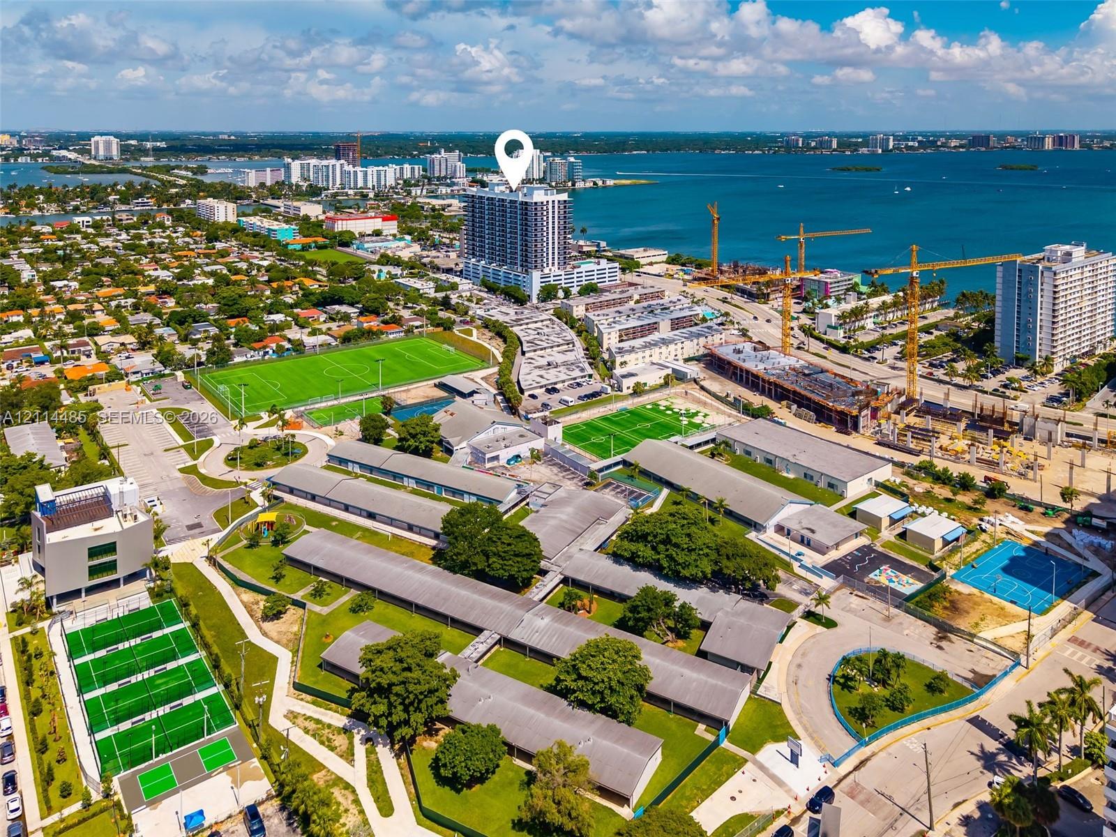 Aerial perspective featuring a waterfront high-rise tower, proximity to multiple recreational turf fields, dedicated hard-court tennis facilities, and established mature canopy trees