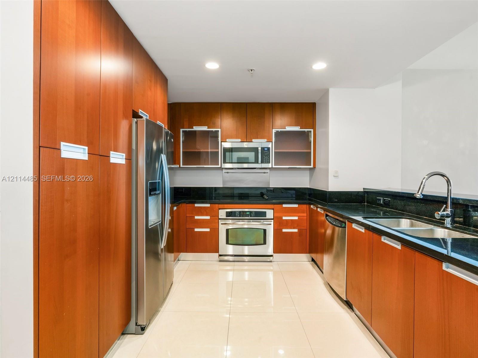 Kitchen featuring custom cabinetry with wood-finish doors, dark stone countertops, stainless steel appliances, and large-format tile flooring