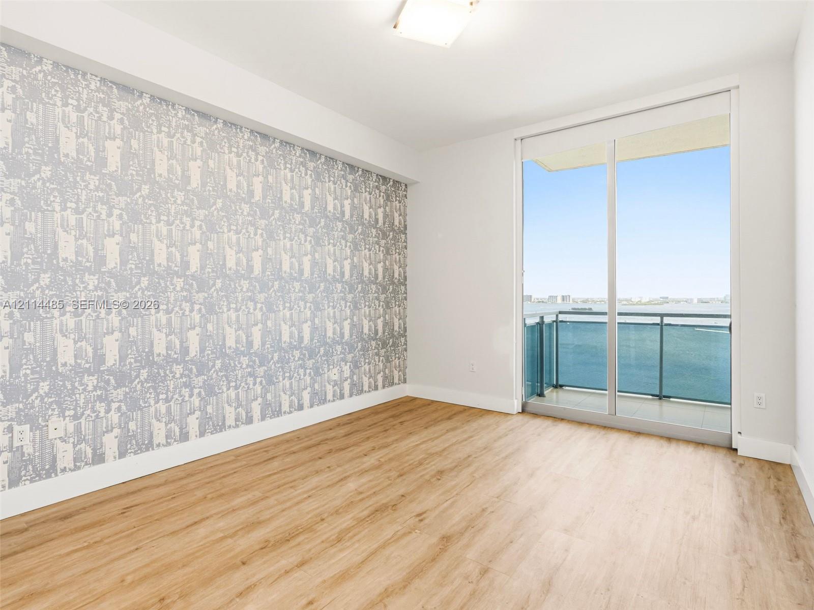 Room featuring wood-finish flooring, a patterned accent wall, a square flush-mount ceiling light, and floor-to-ceiling sliding glass doors opening to a balcony with metal railing