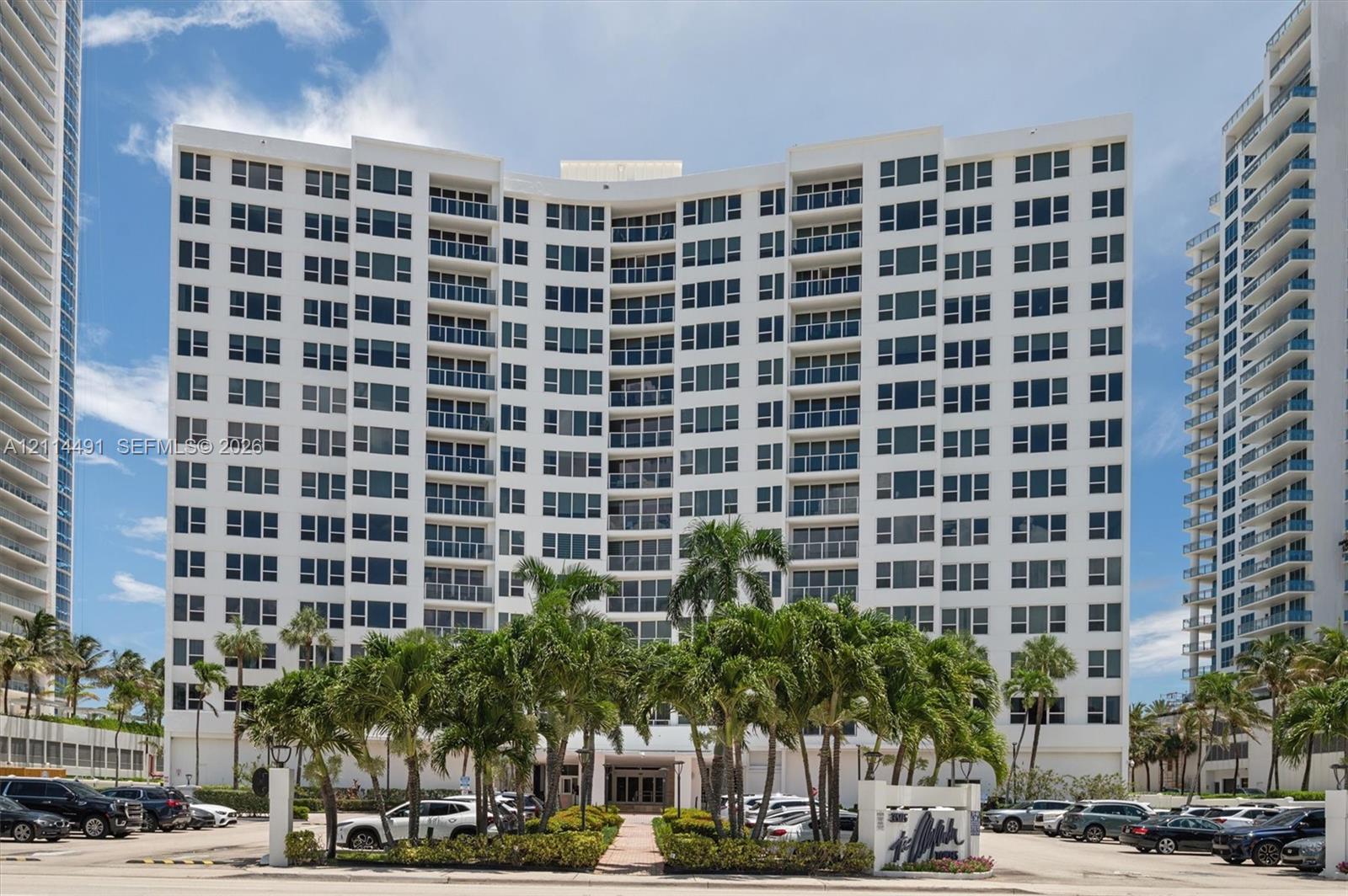 High-rise residential exterior featuring white stucco facade, symmetrical balcony configuration, expansive glass windows, and landscaped grounds with mature palm trees