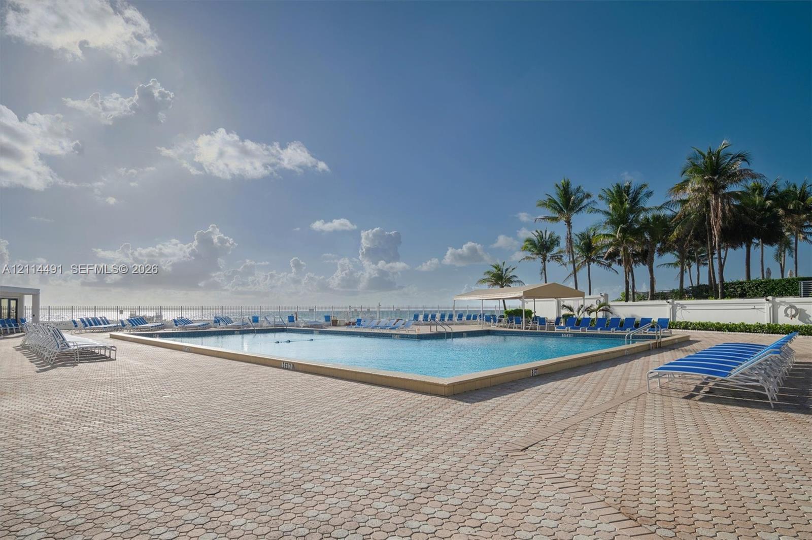 In-ground swimming pool featuring a geometric design, expansive paved patio, permanent shade structure, and perimeter fencing