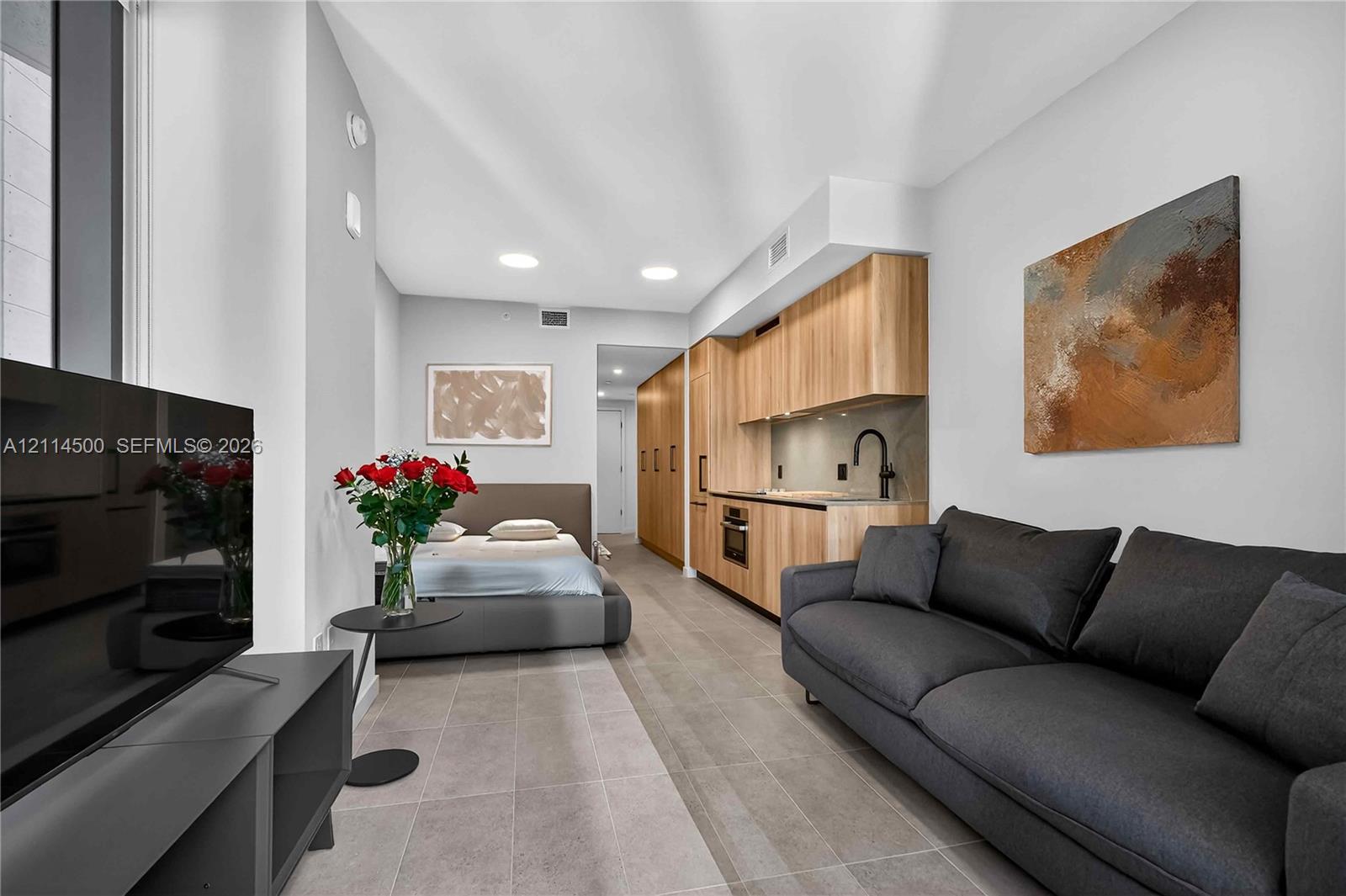 Studio interior featuring wood-finish cabinetry, stone-style tile flooring, recessed ceiling lighting, and a built-in kitchen area with a matte black faucet