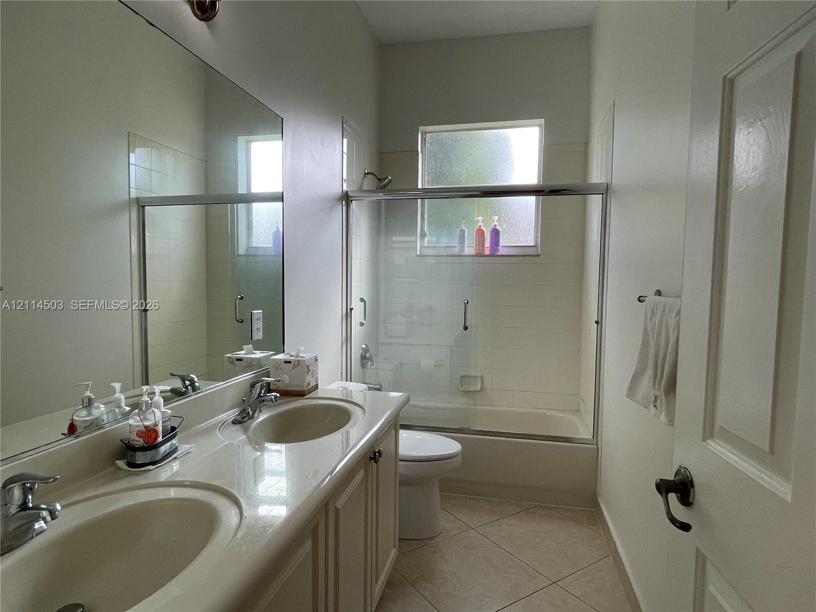 Bathroom featuring a dual-sink vanity with solid-surface countertop, full-width wall mirror, tile flooring, and a tub-shower combination with sliding glass doors