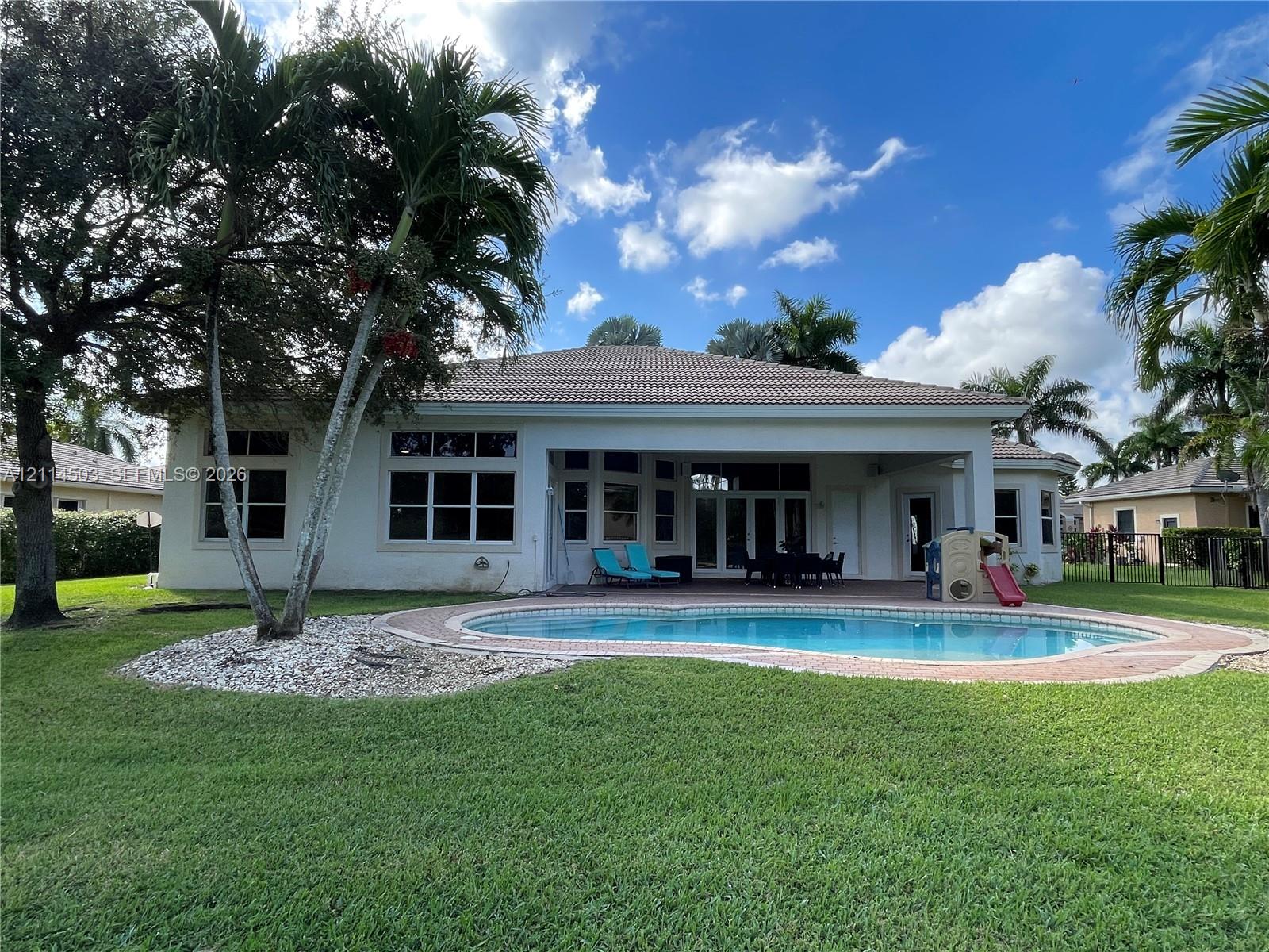 Exterior view featuring a tiled roof, covered patio, curved inground swimming pool with brick paver surround, and mature palm trees