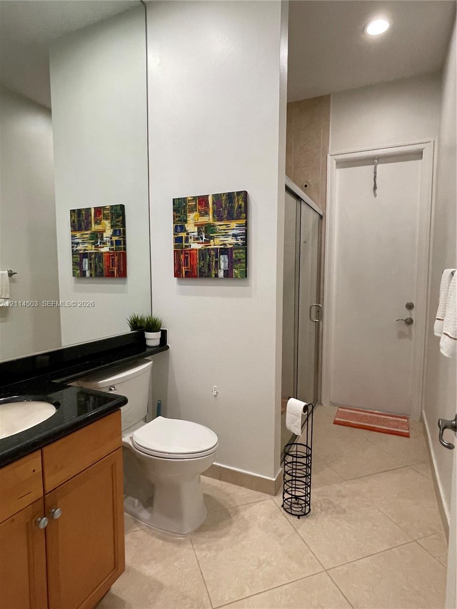 Bathroom featuring a natural wood-finish vanity, dark-toned stone-style countertop with an integrated basin, expansive wall-to-wall mirror, walk-in shower with a glass enclosure, and neutral-toned floor tile laid on a diagonal