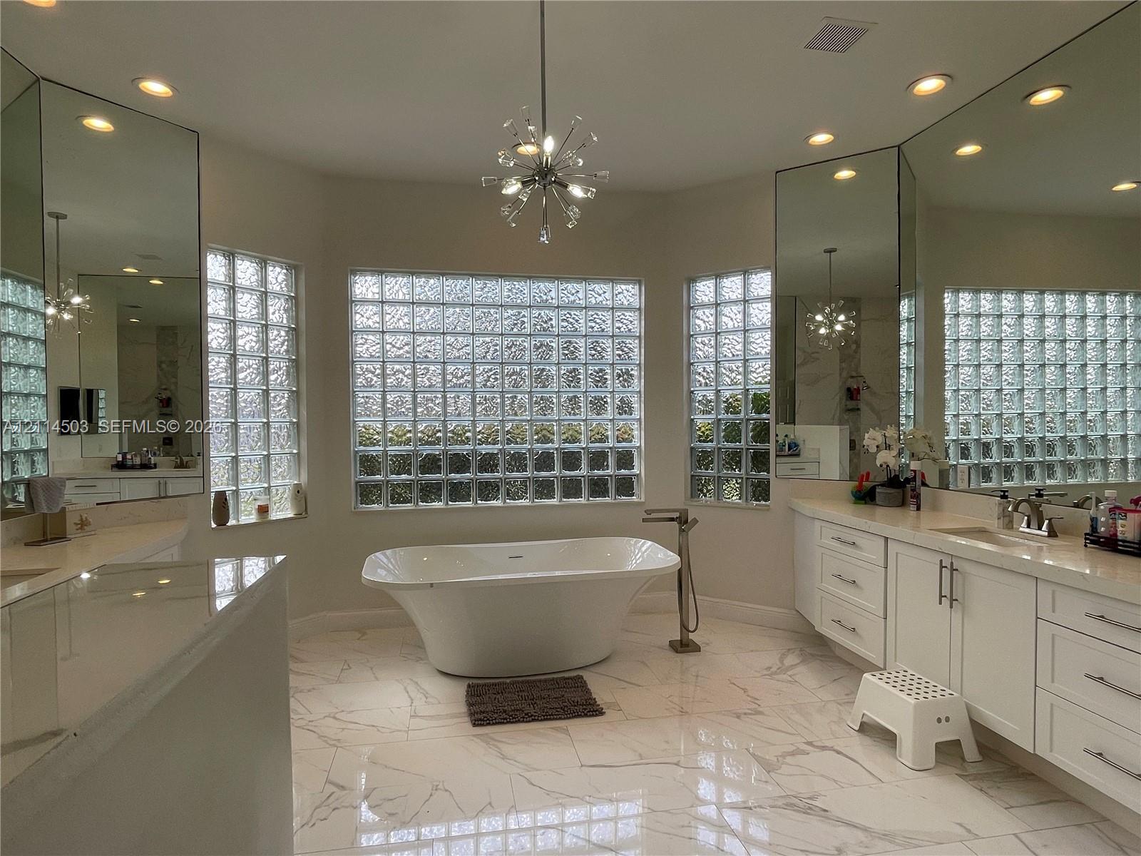 Freestanding soaking tub with floor-mounted faucet, expansive glass block windows, dual white cabinetry, stone-finish tile flooring, and multi-faceted wall mirrors