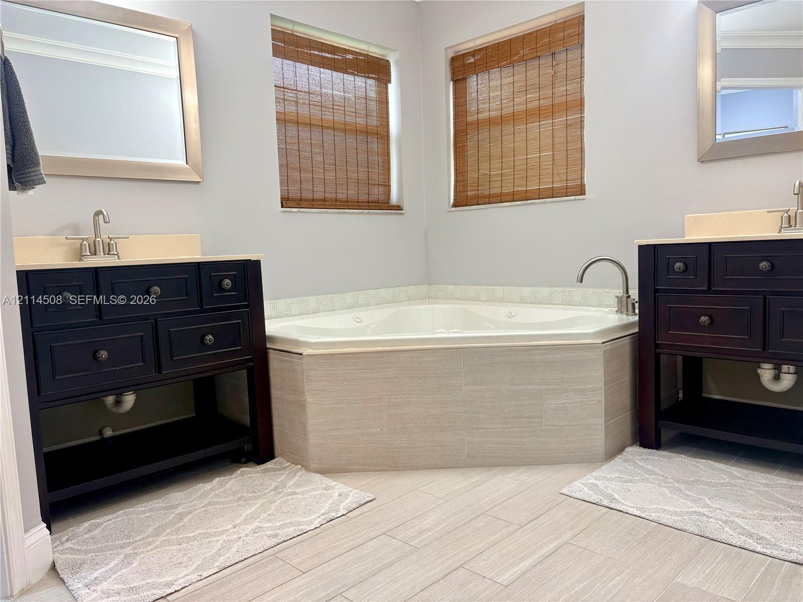 Dual dark-finish wooden vanities with light-toned countertops, corner soaking tub with neutral tile surround, and light-toned tile flooring