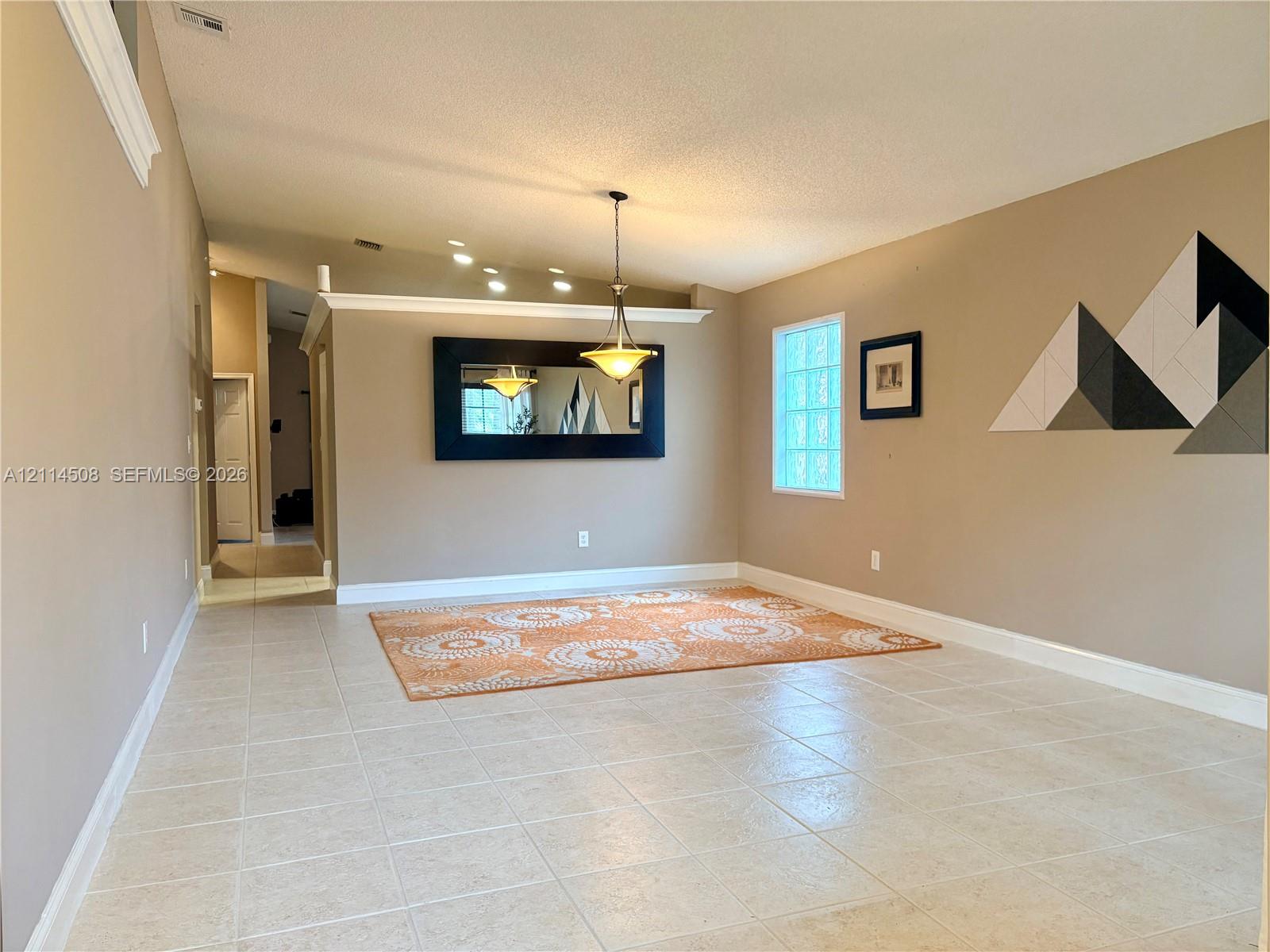 Open-concept interior featuring neutral tile flooring, vaulted ceilings with crown molding, glass block accent window, and a decorative metal pendant light Open-concept interior featuring neutral tile flooring, vaulted ceilings with crown molding, glass block accent window, and a decorative metal pendant light