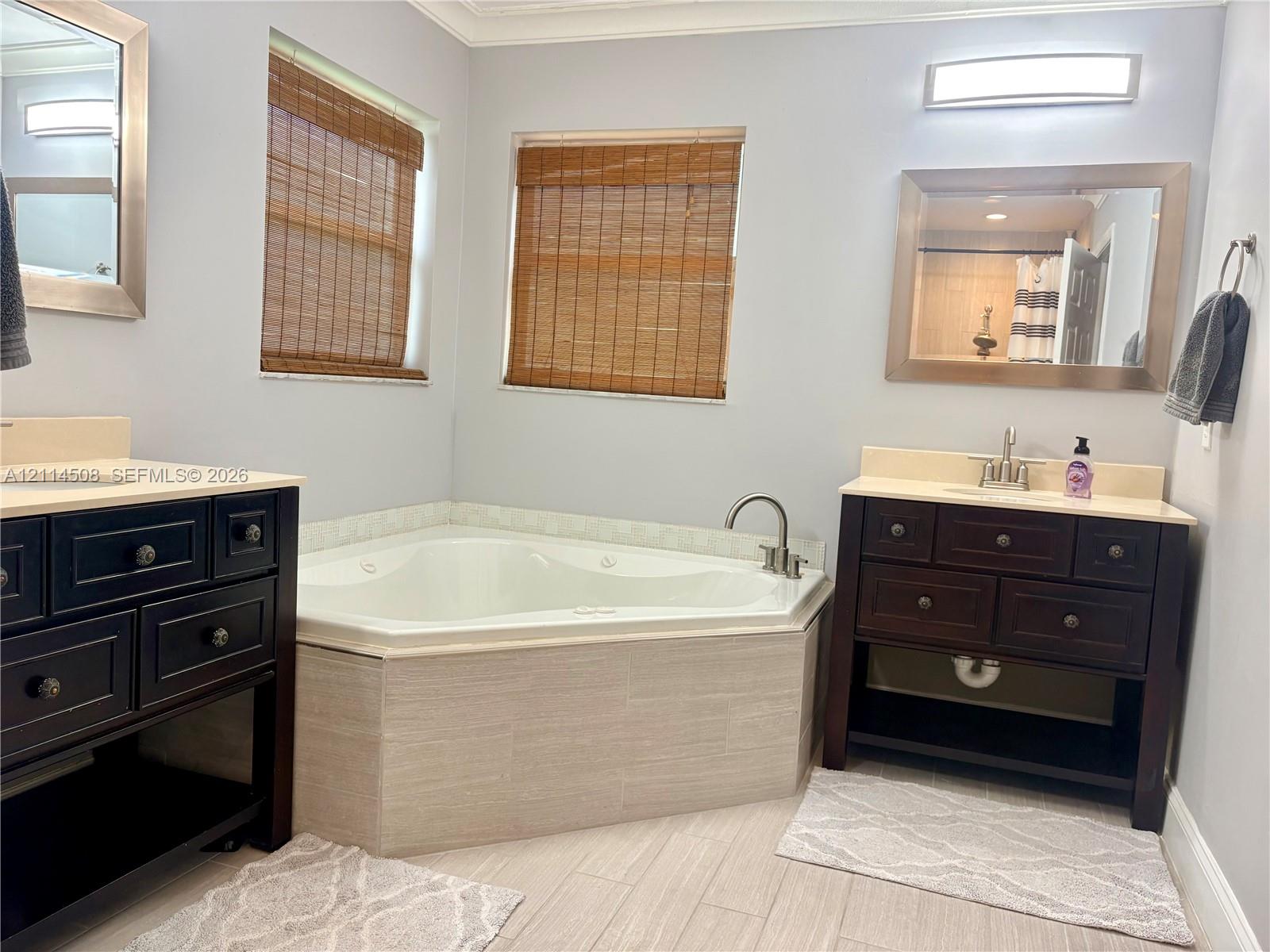 Corner jetted soaking tub with tile surround, dual dark wood vanities with light-toned countertops, wall-mounted light fixtures, and neutral tile flooring