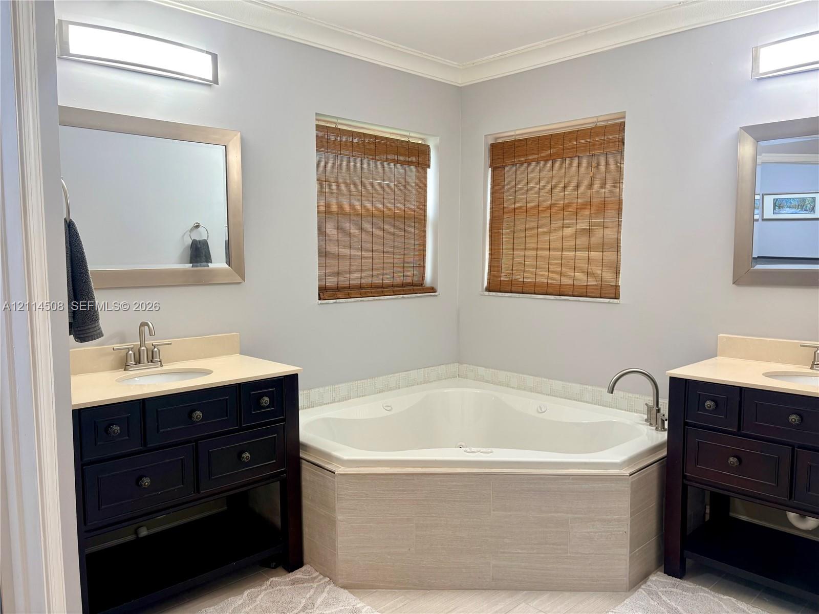 Dual dark wood vanities featuring integrated sinks and stone-surfaced countertops, centered by a built-in corner soaking tub with tile surround and dual wall-mounted light fixtures