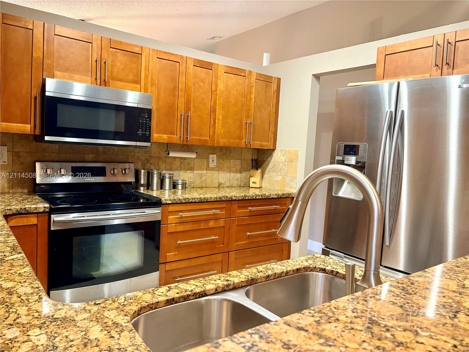 Kitchen featuring natural-finish wood cabinetry, stone-surface countertops, stainless steel appliances, stone tile backsplash, and a dual-basin undermount sink with a high-arc pull-down faucet
