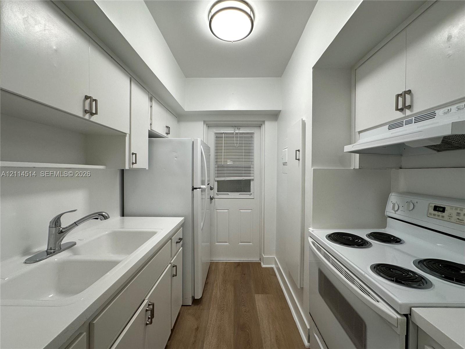 Galley kitchen featuring white cabinetry, dual-basin sink with chrome faucet, integrated range hood, white electric range, and wood-finish flooring Galley kitchen featuring white cabinetry, dual-basin sink with chrome faucet, integrated range hood, white electric range, and wood-finish flooring