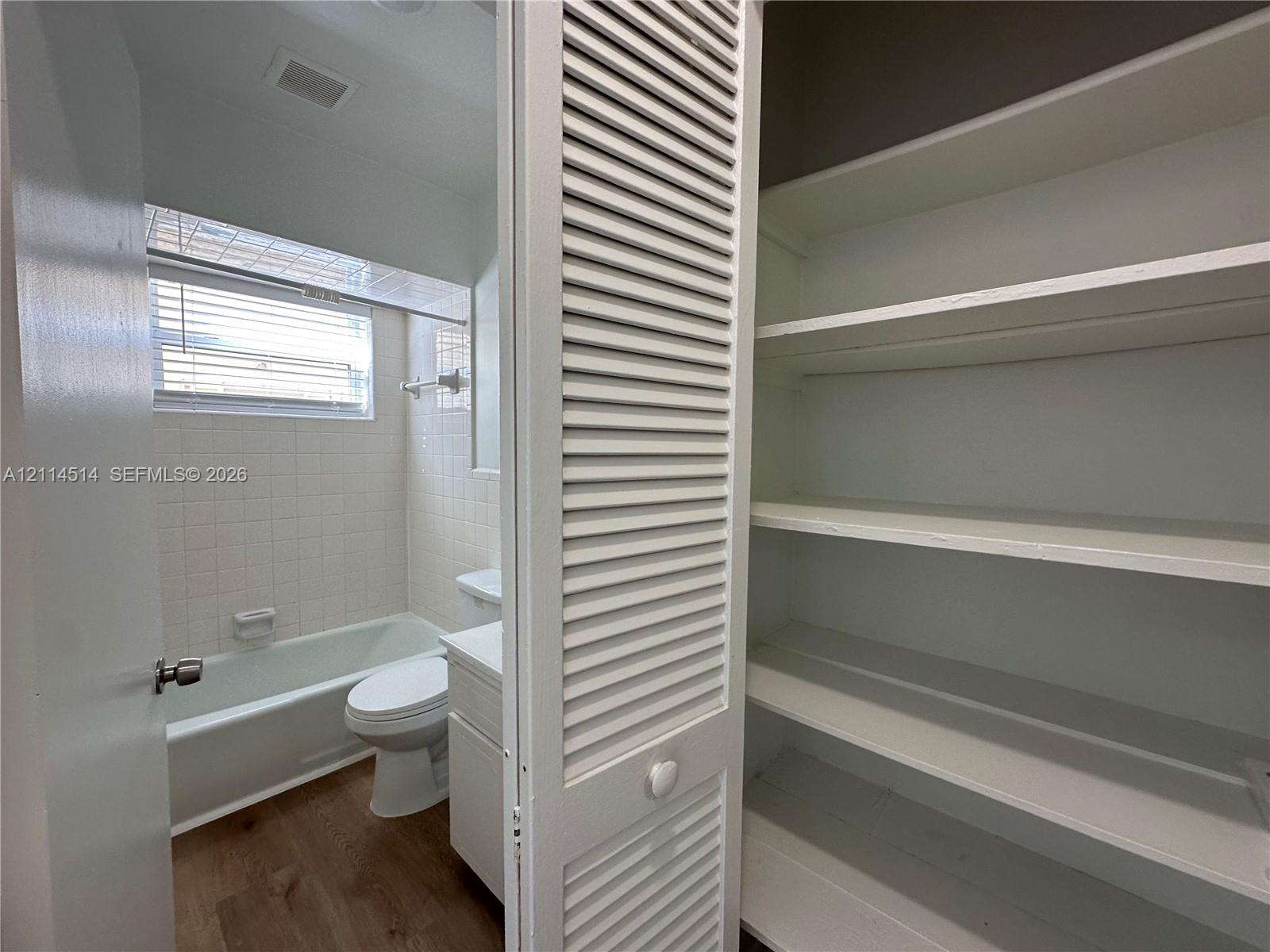 Bathroom featuring a built-in linen closet with louvered doors, white tiled walls, a bathtub with a shower surround, a white vanity, and wood-finish flooring Bathroom featuring a built-in linen closet with louvered doors, white tiled walls, a bathtub with a shower surround, a white vanity, and wood-finish flooring