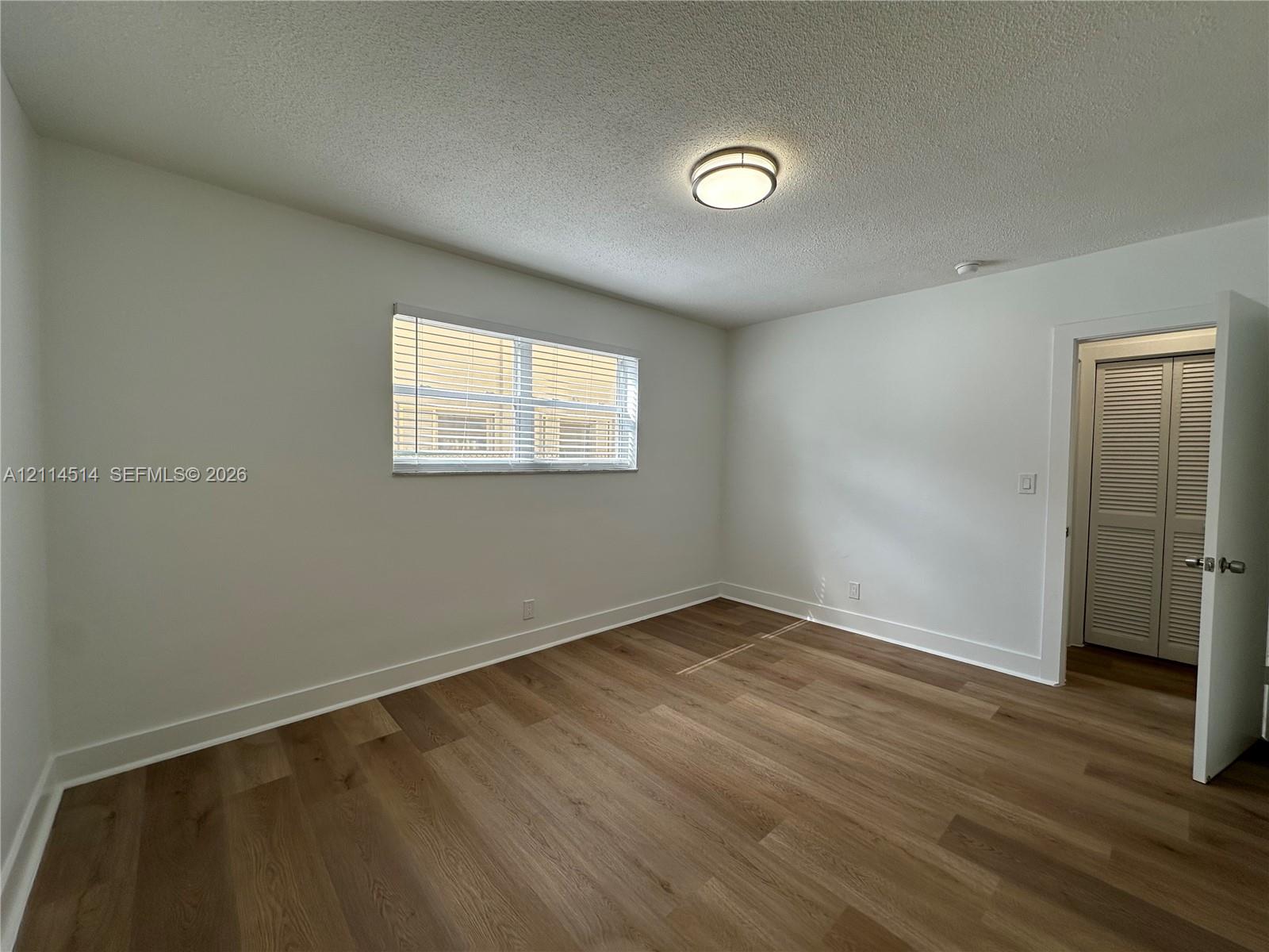 Interior room featuring wood-finish flooring, a neutral color palette, a central flush-mount ceiling light, a horizontal window with slatted blinds, and a closet with louvered double doors