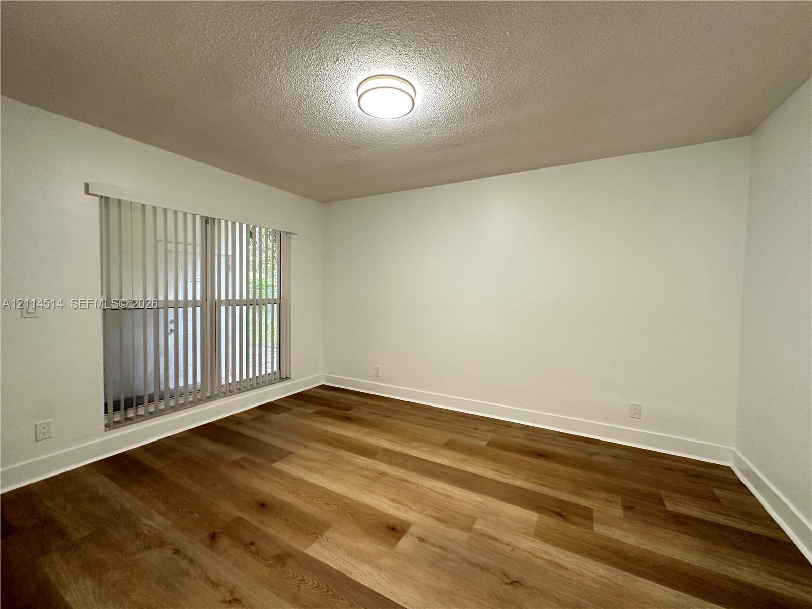 Neutral interior space featuring wood-finish flooring, white walls, vertical blinds, and a central flush-mount ceiling light Neutral interior space featuring wood-finish flooring, white walls, vertical blinds, and a central flush-mount ceiling light