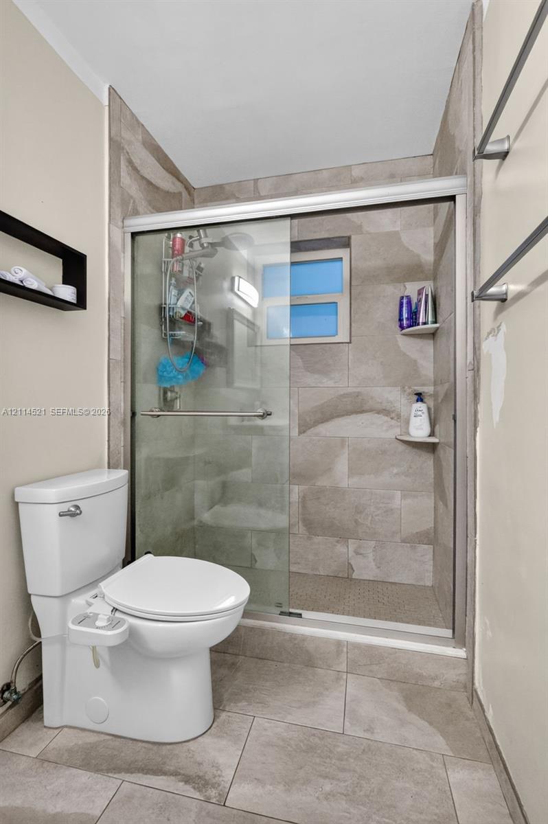 Walk-in shower with sliding glass door, neutral-toned stone-style wall tile, integrated corner shelving, and large-format floor tile