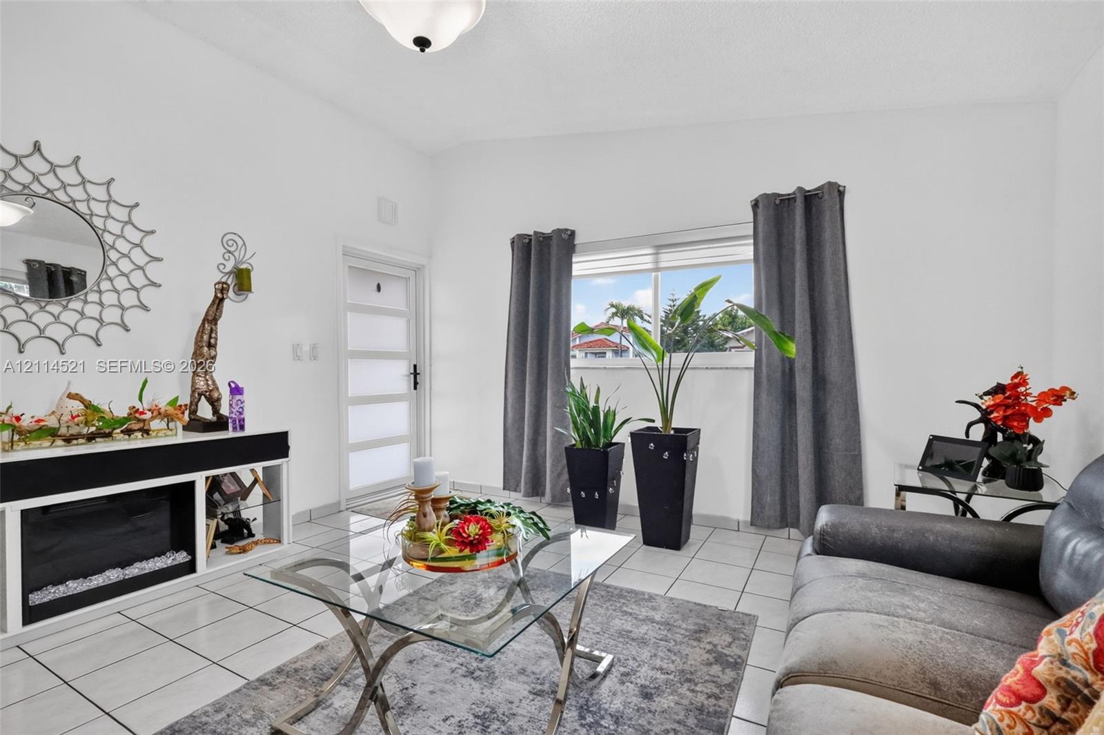 Living space featuring white ceramic tile flooring, a horizontal paneled entry door with frosted glass inserts, a built-in media console, and a large window with blind treatments Living space featuring white ceramic tile flooring, a horizontal paneled entry door with frosted glass inserts, a built-in media console, and a large window with blind treatments