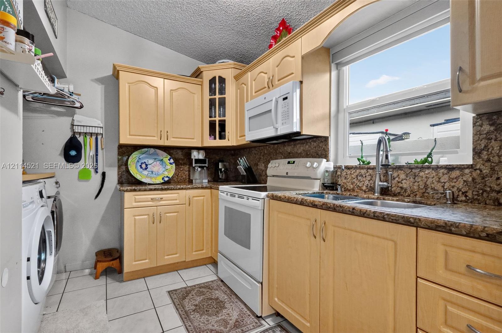 Kitchen featuring natural-finish wood cabinetry, stone-patterned countertops, a double-basin stainless steel sink with a goose-neck faucet, and white tile flooring Kitchen featuring natural-finish wood cabinetry, stone-patterned countertops, a double-basin stainless steel sink with a goose-neck faucet, and white tile flooring