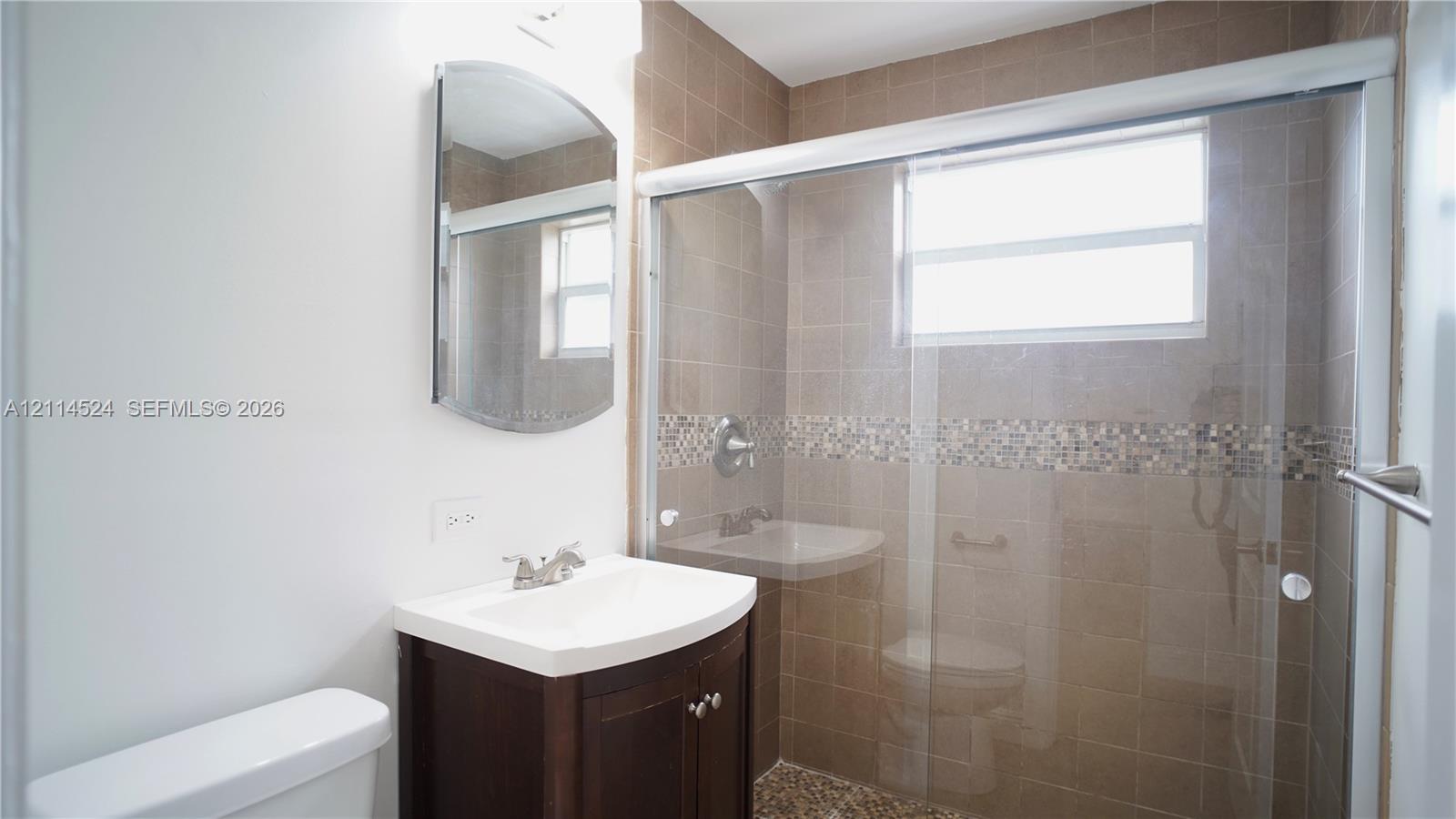 Bathroom featuring a dark wood-finish vanity with a white basin, a wall-mounted arched mirror, and a walk-in shower with tiled surround, a decorative mosaic tile accent border, and a glass sliding door