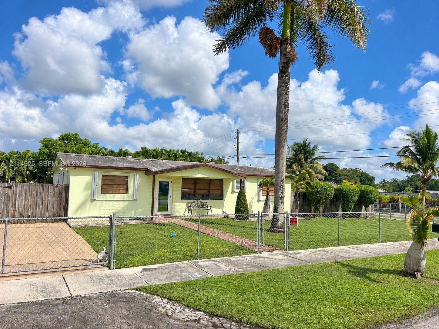 Single-story exterior featuring a brick walkway, chain-link perimeter fencing, mature palm trees, and a paved side driveway Single-story exterior featuring a brick walkway, chain-link perimeter fencing, mature palm trees, and a paved side driveway