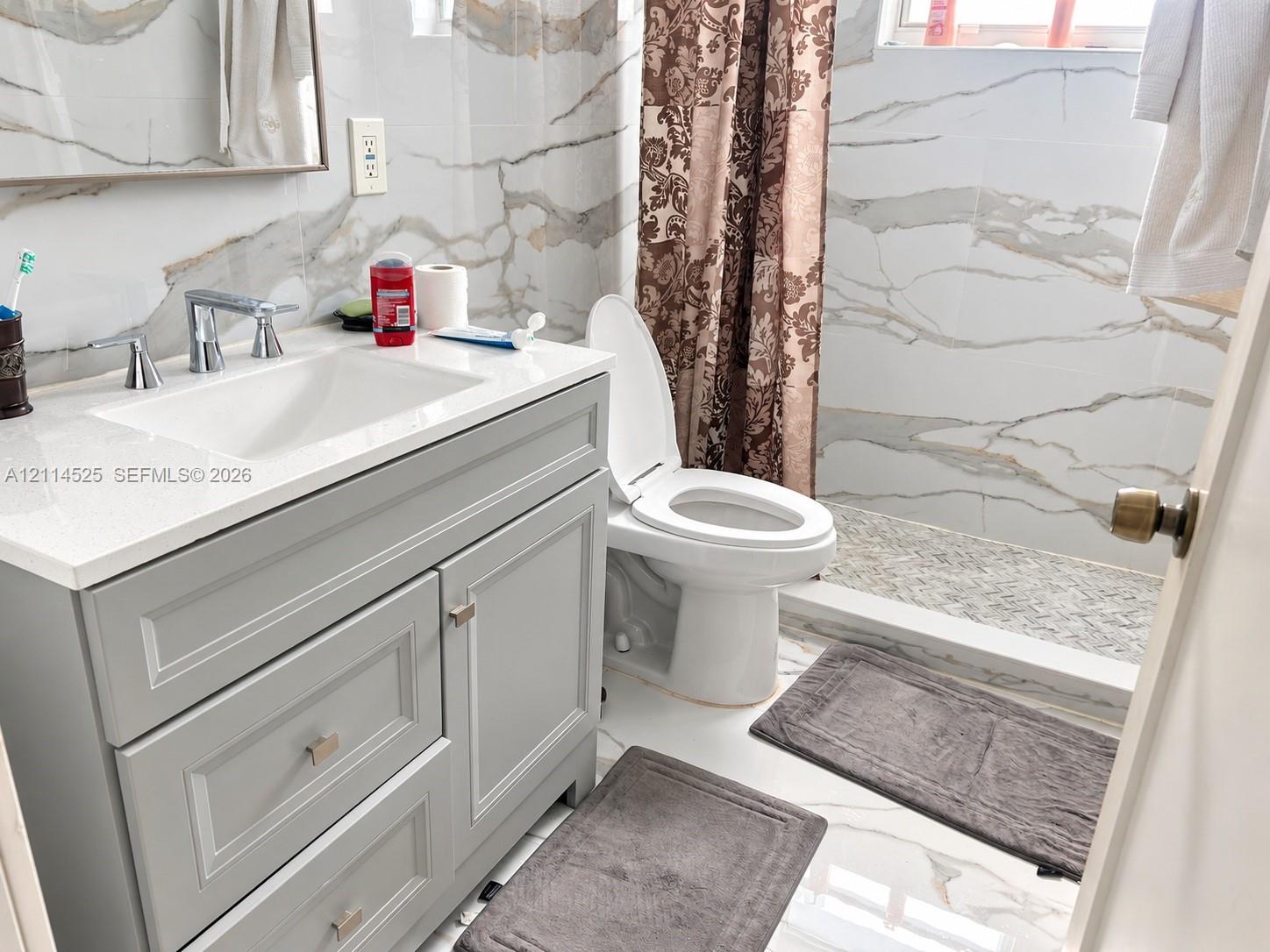 Bathroom featuring a grey shaker-style vanity with a solid surface countertop, an undermount sink with a polished chrome faucet, and large-format wall tiling with a vein pattern