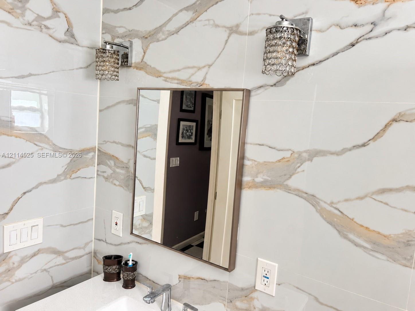 Large-format wall tiling with gold and gray veining, square mirror with metallic frame, and dual crystal-accented wall sconces