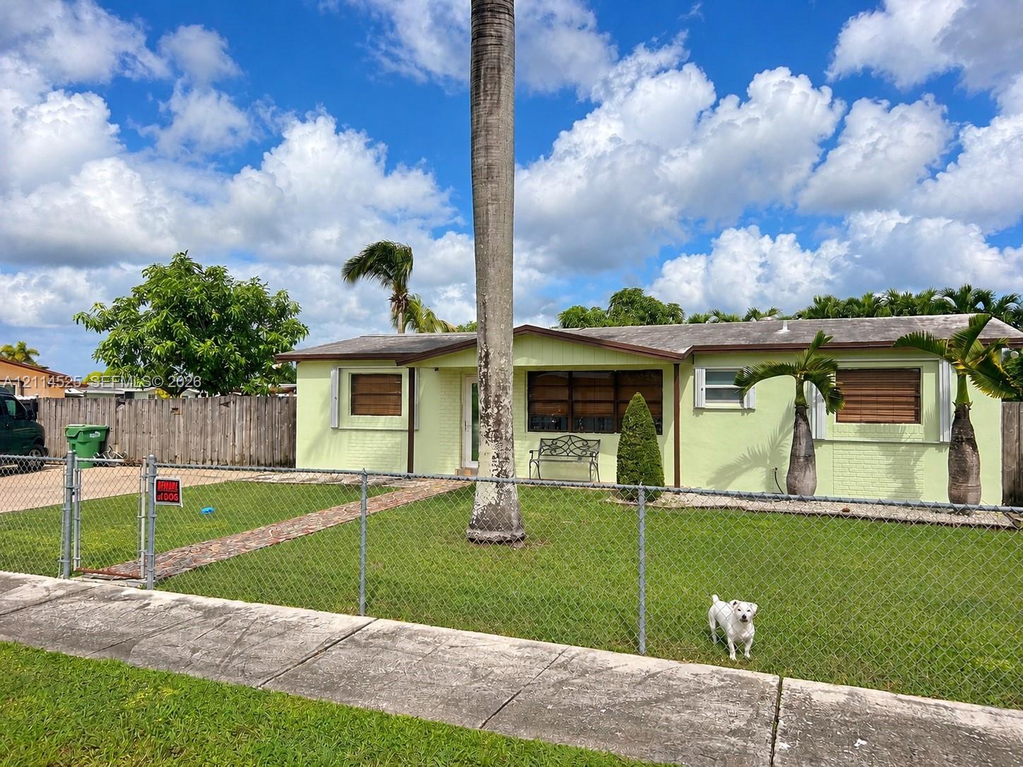 Single-story exterior featuring painted masonry siding, a gabled roofline, metal storm shutters, and a chain-link fenced front yard with mature landscaping Single-story exterior featuring painted masonry siding, a gabled roofline, metal storm shutters, and a chain-link fenced front yard with mature landscaping