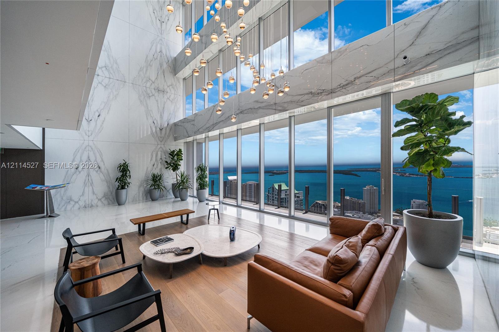 Double-height ceilings with floor-to-ceiling glass windows, full-wall stone cladding, integrated multi-pendant lighting fixture, and light-tone wood-finish flooring