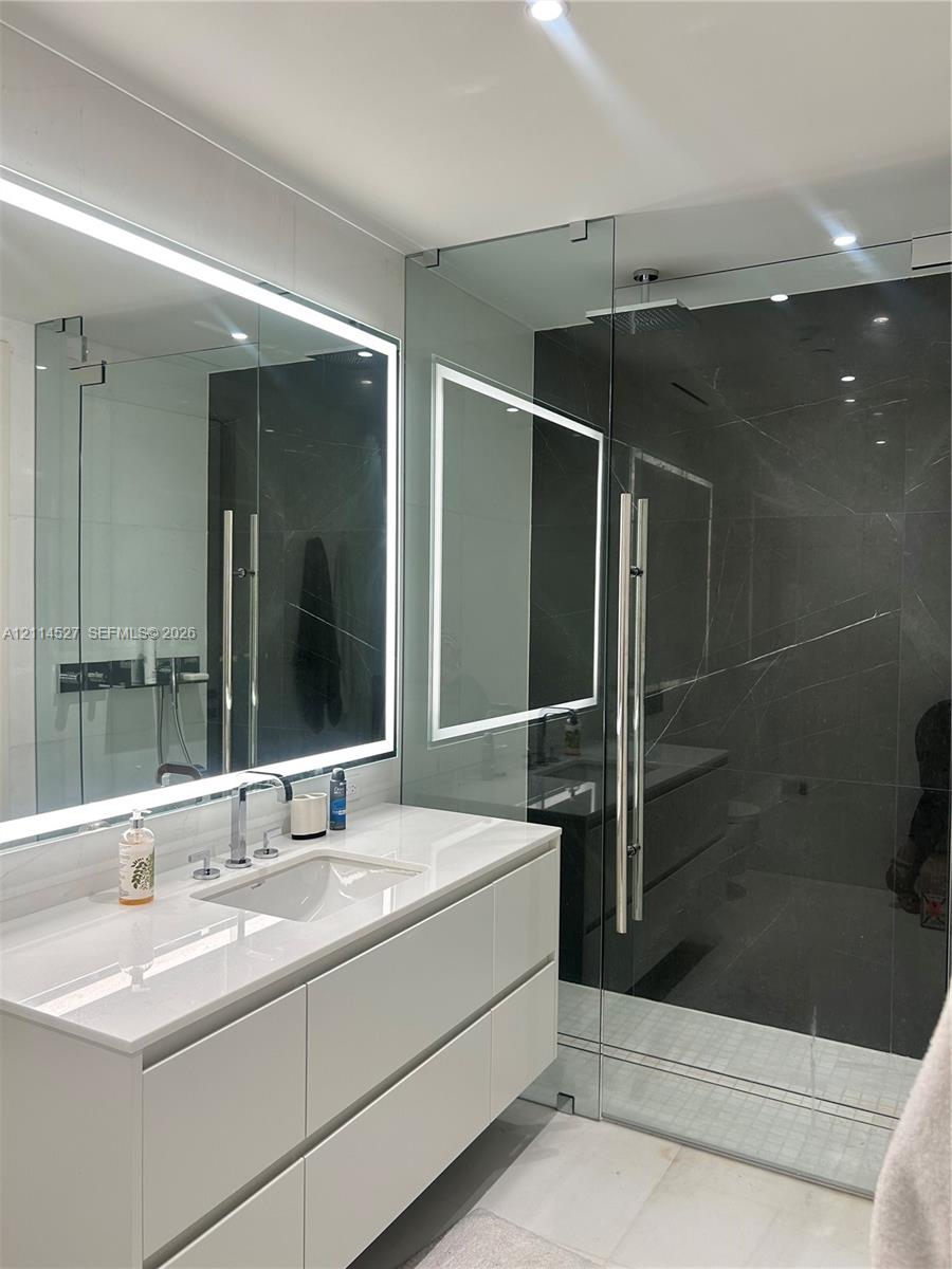 Wall-mounted floating vanity with integrated rectangular sink and stone-surface countertop, frameless glass-enclosed shower featuring large-format dark tile, and integrated LED-backlit mirror