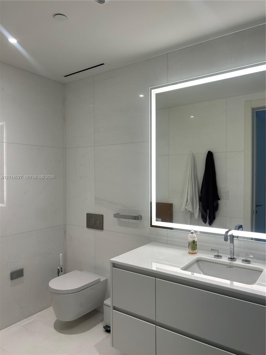 Wall-mounted toilet with a dual-flush actuator plate, large-format tile wall cladding, modern floating vanity with a seamless countertop, rectangular undermount sink, and integrated LED-backlit mirror