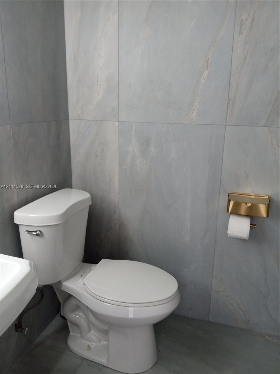 Full-height large-format wall and floor tiling with subtle veining, dual-flush toilet, and metallic gold-tone toilet paper dispenser