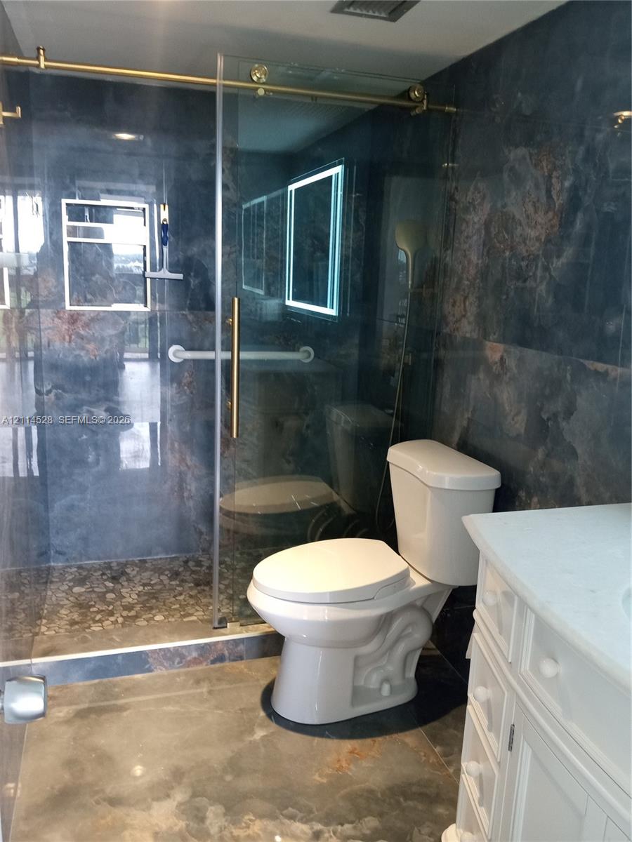 Walk-in shower featuring gold-tone hardware, a glass sliding door, and a pebble-stone floor, complemented by dark-toned tile walls, a vanity with a light-toned countertop, and a wall-mounted light-up mirror