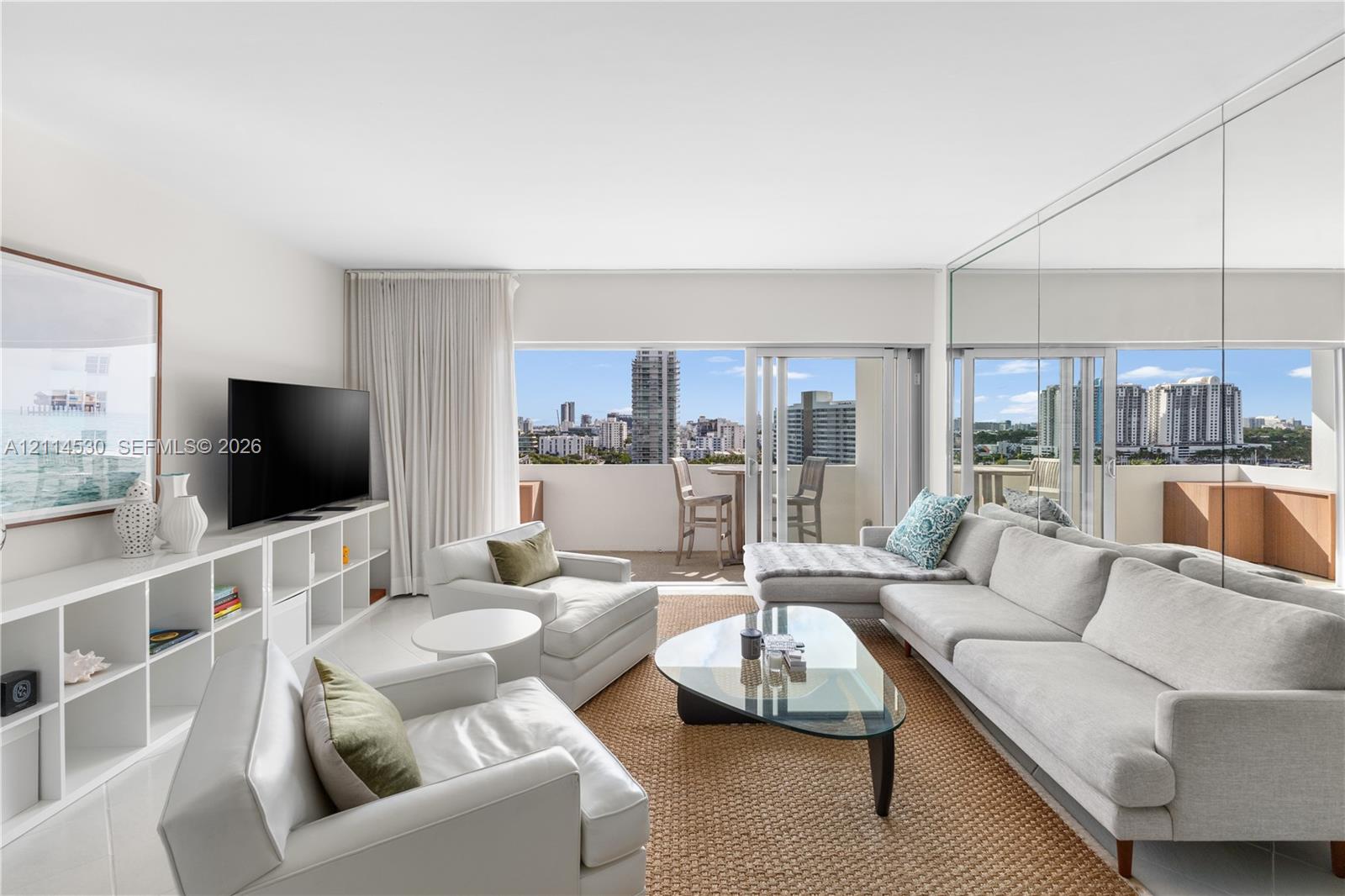 Living area featuring floor-to-ceiling mirrored walls, custom built-in white shelving, sliding glass balcony doors, and panoramic city skyline scenery Living area featuring floor-to-ceiling mirrored walls, custom built-in white shelving, sliding glass balcony doors, and panoramic city skyline scenery