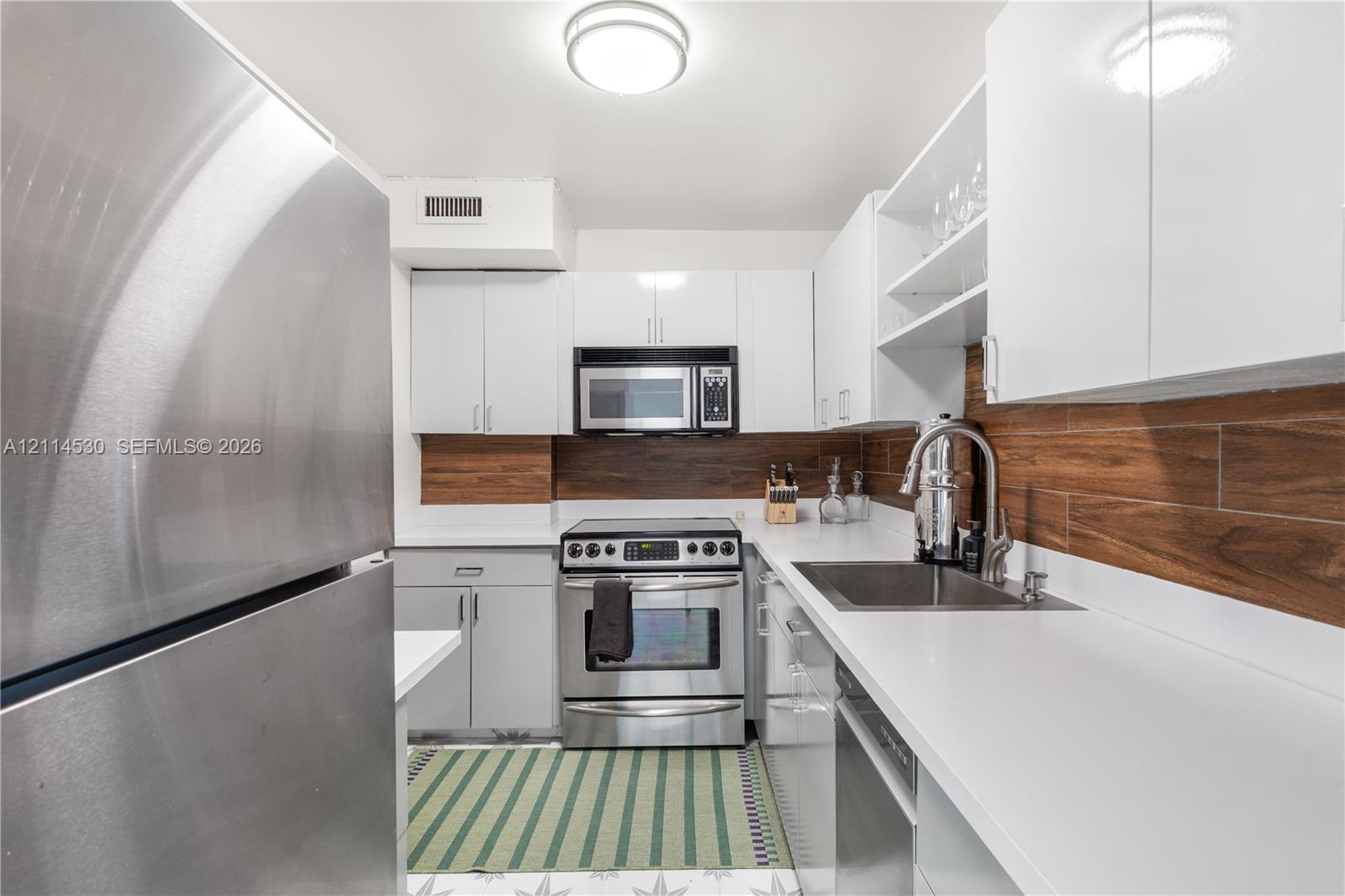 Kitchen featuring stainless steel appliances, white gloss cabinetry, solid surface countertops, and wood-finish tile backsplash
