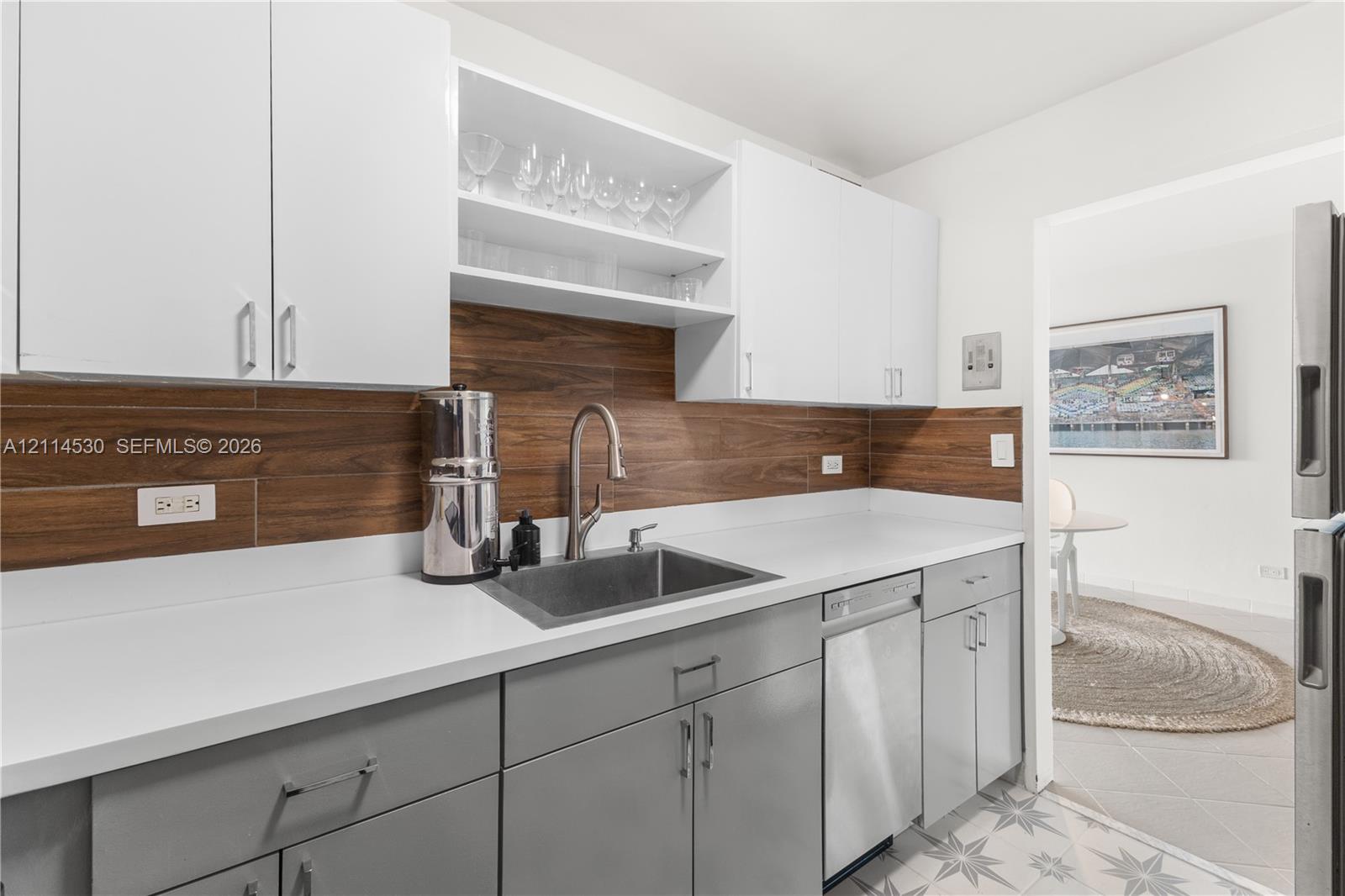 Kitchen featuring white upper cabinetry, grey lower cabinetry, wood-tone backsplash, solid-surface countertops, and an undermount sink with a high-arch faucet