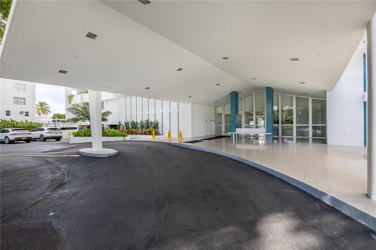 Covered porte-cochre entry featuring recessed lighting, a paved drop-off area, blue decorative columns, and full-height glass storefront glazing