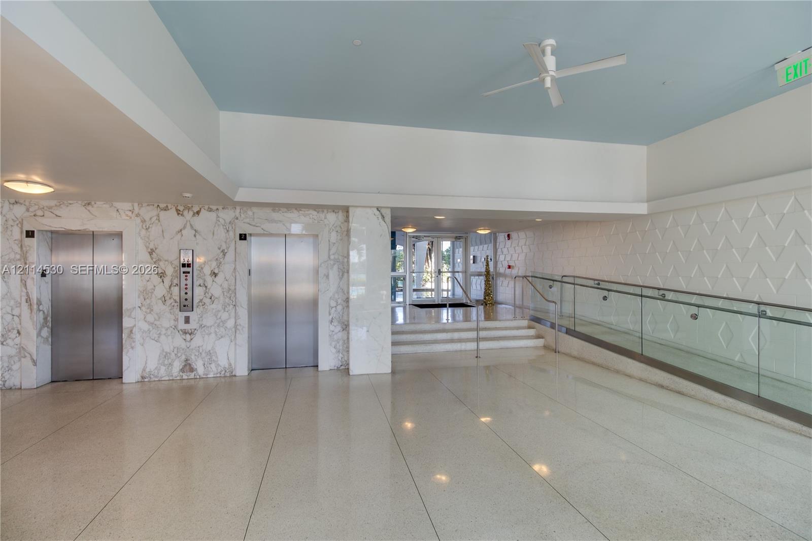 Lobby area featuring dual stainless-steel elevators, stone-paneled accent walls, polished stone flooring, a glass-panel ramp railing, and textured wall detailing