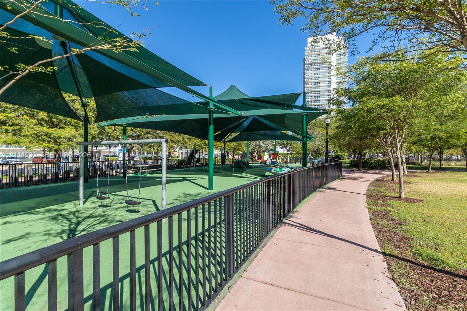 Recreational space featuring a synthetic turf surface, metal perimeter fencing, elevated sunshades, and a paved walkway