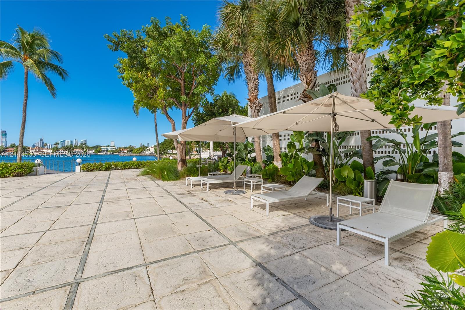 Waterfront patio featuring expansive stone tile pavers, mature palm trees, lush tropical landscaping, and a decorative concrete masonry wall