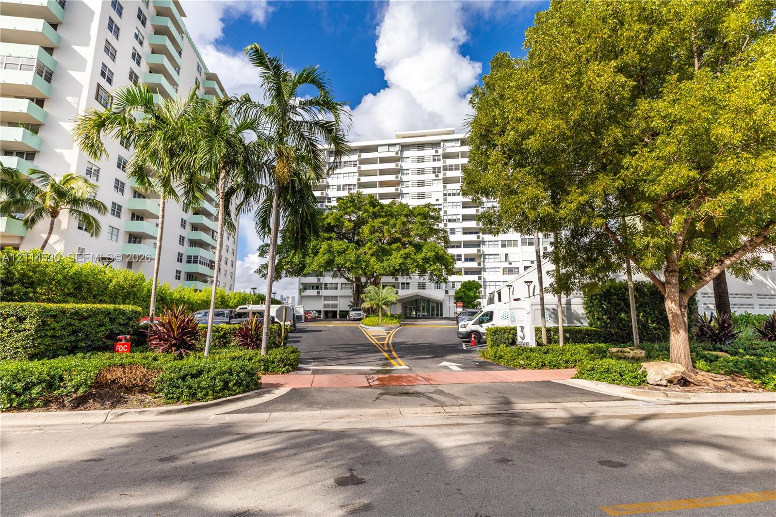 High-rise residential exterior featuring white balconies, asphalt driveways, mature landscaping, and palm trees