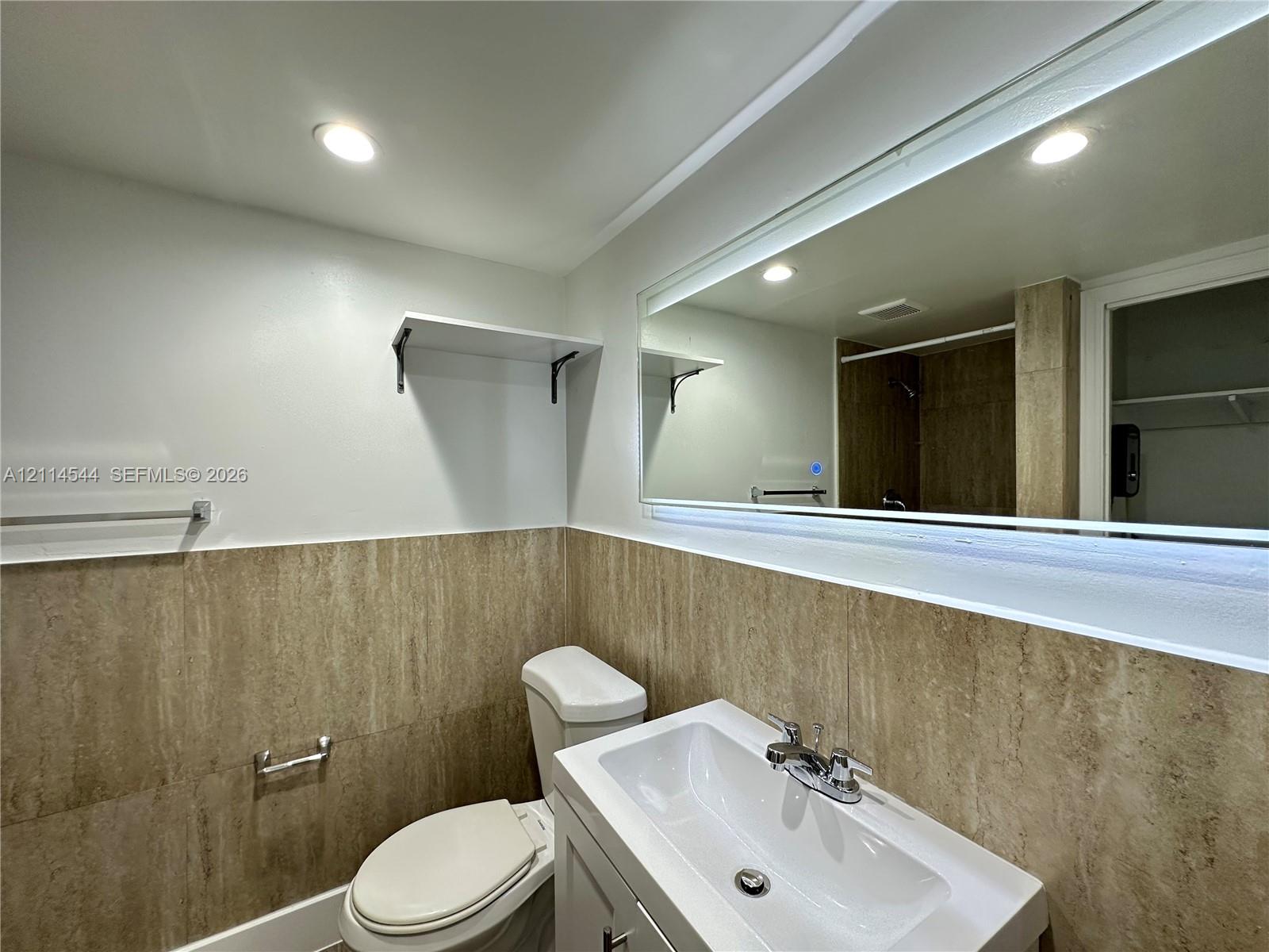 Bathroom featuring floor-to-ceiling stone-tile wainscoting, a vanity with an integrated basin, a polished chrome faucet, and a large wall-mounted mirror with integrated illumination