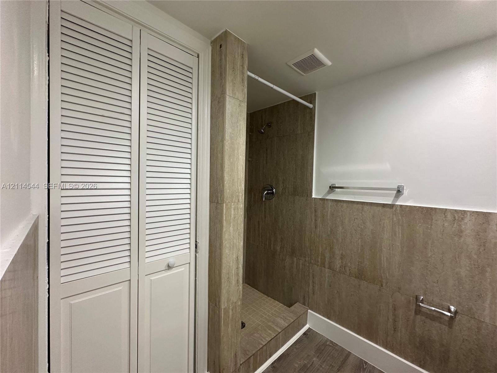 Walk-in shower featuring large-format stone-textured tile, white louvered-door closet, metallic hardware, and wood-finish flooring